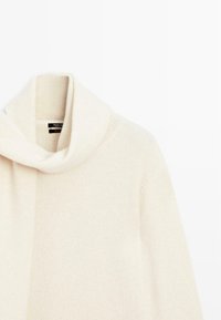 Pullover in cashmere color crema con colletto a sciarpa drappeggiato appeso su uno sfondo bianco.