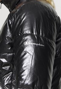 Chaqueta de plumas negra hecha de un material brillante y reflectante. Presenta un logo plateado en la manga y un diseño de cuello alto.