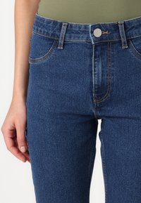 Jean en denim bleu taille haute avec une texture lisse, des coutures contrastées, deux poches avant et une fermeture à bouton à la taille.