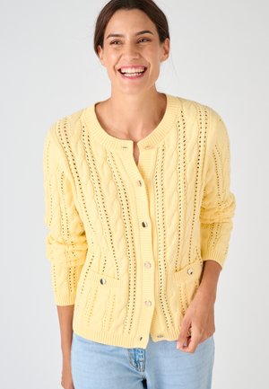 Femme souriante portant un cardigan tricoté câble jaune pâle avec des boutons argentés et des poches devant, associé à un jean bleu clair, debout devant un fond blanc.