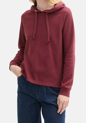 Personne portant un sweat à capuche bordeaux avec des cordons et un jean bleu, main gauche dans la poche, montrée des épaules jusqu'au haut des cuisses sur un fond uni.
