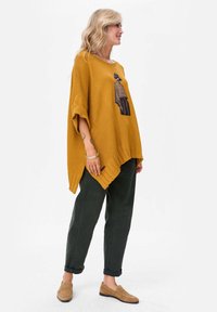 Musztardowo żółty, oversize'owy ponczo o fakturze prążkowanej z bocznymi rozcięciami, ozdobiony czarnym nadrukiem graficznym. W zestawie z ciemnozielonymi spodniami i jasnobrązowymi mokasynami.