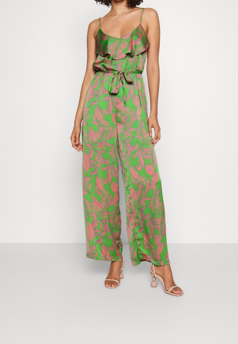 Femme portant une combinaison pantalon large avec un motif de feuilles roses sur fond vert et des bretelles fines, associée à des sandales à talons hauts couleur nude.
