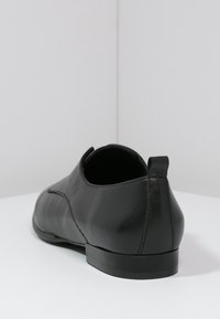 Czarna skórzana broszka o okrągłym kształcie, gładkiej teksturze i lekko wyższym obcasie. Posiada pętelkę z tyłu, co ułatwia zakładanie i ściąganie.