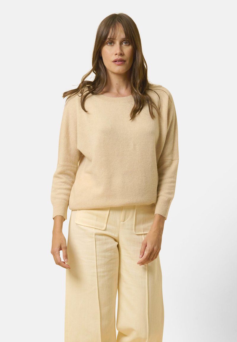 Beiger gestrickter Pullover mit weitem Ausschnitt und langen Ärmeln, kombiniert mit hellgelben, weit geschnittenen Hosen mit großen Taschen.