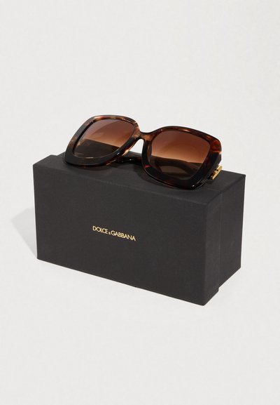 Καφετιά ηλιόφωνα γυαλιά υπερμεγέθη με φακούς διαβάθμισης, που ακουμπούν σε μαύρο κουτί με χρυσά γράμματα "DOLCE & GABBANA".