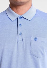 Polo bleu clair avec un col texturé et à motifs, trois boutons bleus et un logo brodé sur la poche poitrine gauche.
