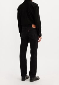 Personne portant une chemise noire à manches longues, un jean noir droit avec un patch en cuir, et des chaussures noires, debout dos à la caméra.
