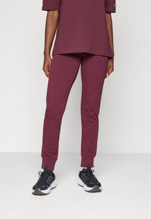 Pantalons de survêtement bordeaux en matériau doux, avec une coupe fuselée et des poignets élastiques. Associés à des chaussures de sport noires avec des accents blancs.