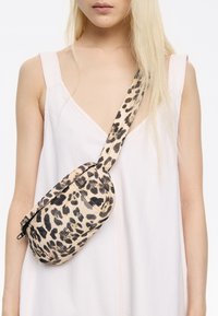 Torebka typu crossbody z materiału w panterkę, z czarnymi akcentami, w zaokrąglonym kształcie oraz regulowanym paskiem, noszona na jasnoróżowej sukience.