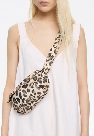 Torebka typu crossbody z materiału w panterkę, z czarnymi akcentami, w zaokrąglonym kształcie oraz regulowanym paskiem, noszona na jasnoróżowej sukience.