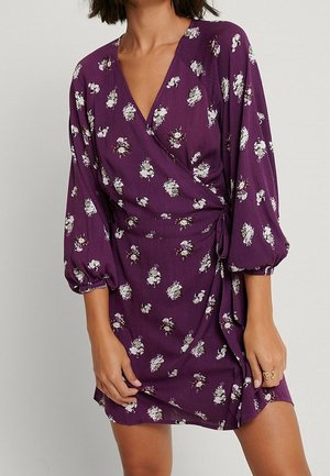 Mujer que lleva un vestido cruzado morado con estampados florales blancos y mangas abullonadas hasta el codo, de pie frente a un fondo liso.
