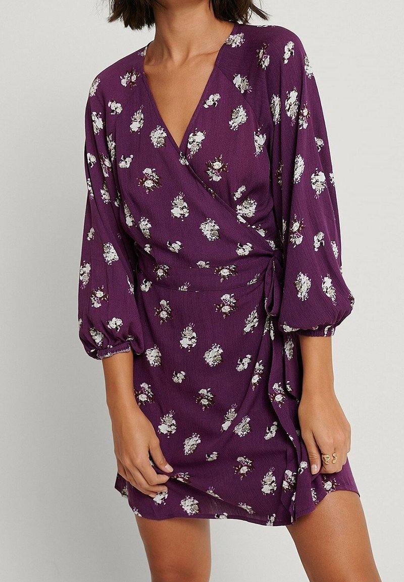 Femme portant une robe portefeuille violette avec des imprimés floraux blancs et des manches bouffantes trois-quarts, debout devant un fond uni.