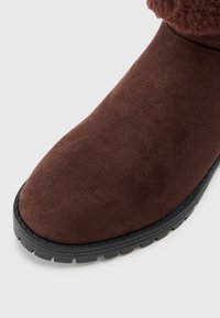 Botine din suede maro, cu vârf rotunjit și accent texturat, având o talpă din cauciuc negru cu șanțuri pentru tracțiune.