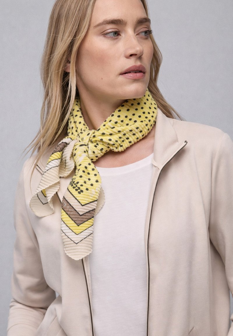 Femme portant une veste beige clair et une chemise blanche, avec une écharpe jaune à pois et motif chevron nouée autour du cou.