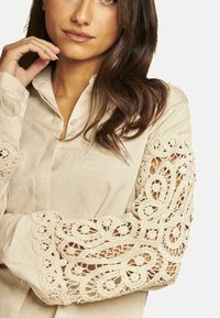 Blusa de lino color beige claro con intrincados detalles de encaje crema en las mangas, que presentan espirales y patrones florales.