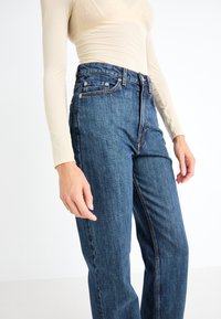 Kõrge vöökohaga sinised denim püksid sirge jala disainiga, ees on taskud ja vastandõmblused. Kanga tekstuurne viimistlus.
