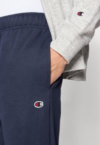 Pantalon de survêtement bleu marine avec taille élastique et poches latérales, arborant un logo brodé blanc et rouge. Complété par un sweat-shirt gris clair.