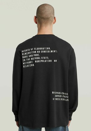 Mann trägt ein schwarzes Langarmshirt mit weißem Text auf dem Rücken: "Ohne Ausschmückung, Verminderung oder Verbergung; nackig und rein."