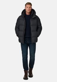 Svart vadderad jacka med huva och quiltad design. Bärs över en mörkblå tröja, tillsammans med mörka jeans och bruna snörkängor.