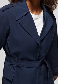 Kobieta w granatowym płaszczu trencz z paskiem, na białej koszulce z okrągłym dekoltem, z częściowo widocznymi kręconymi włosami.