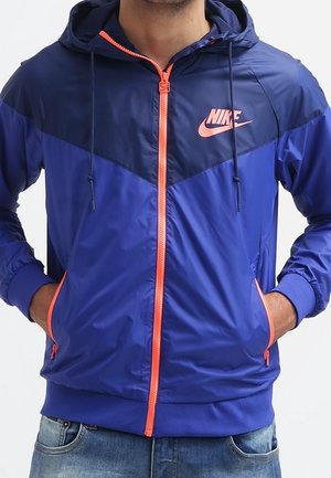 Mężczyzna w niebieskiej kurtce z kapturem Nike z pomarańczowym suwakiem i logo, z rękami w kieszeniach, ubrany w niebieskie dżinsy.