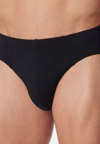Nahaufnahme einer Person, die schlichte schwarze Herrenunterwäsche trägt, wobei der Bund und die Oberschenkel gegen einen neutralen Hintergrund zu sehen sind.