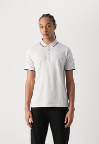 Weißes Poloshirt mit Kragen, mit dunkelblauen Abschlüssen am Kragen und an den Ärmeln. Weicher Stoff, Knopfverschluss und Logo auf der Brust gestickt.