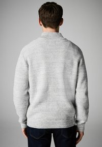 Suéter de punto gris con cuello alto y dobladillo acanalado. Patrón texturizado con ligeras variaciones en la tonalidad. Llevado sobre unos jeans azul oscuro.