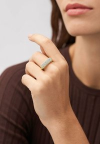 Guldfarvet rektangulært ring med en struktureret overflade, sat med flere klare sten, båret på en hånd med en naturlig hudtone.