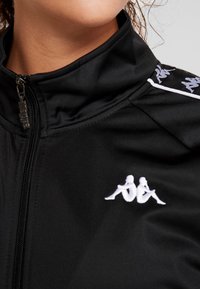 Gros plan d'une veste noire à fermeture éclair avec un logo blanc représentant deux figures assises dos à dos sur la poitrine et répété sur l'épaule.