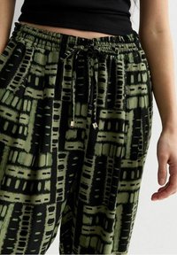 Vihreät ja mustat kuviolliset housut, joissa on joustava vyötärönauha ja kiristysnyöri. Housuissa on geometrisia kuvioita ja rento istuvuus. Lähikuva rakenteellisesta kankaasta.