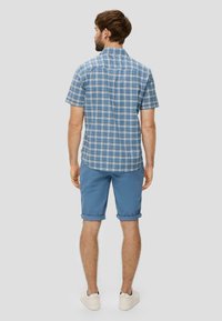 Korteblouw geruit shirt met korte mouwen in blauw en wit, gecombineerd met lichtblauwe opgerolde shorts. Model wordt van achteren getoond, draagt witte sneakers.