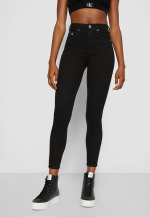 Jeans negros ajustados de cintura alta con una textura suave, que cuentan con cuatro bolsillos y herrajes plateados, combinados con zapatillas de caña alta negras.