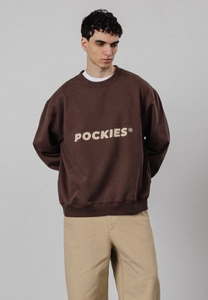 Jonge man met krullend donker haar, dragend een bruine oversized trui met de tekst "POCKIES" en beige broek tegen een effen grijze achtergrond.