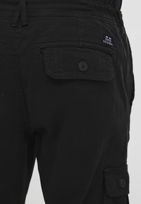 Svart cargo-bukser med teksturert stoff, flere lommer, knappeåpning og et merkeemblem på baksiden.