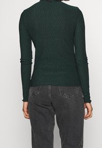 Dunkelgrünes Langarmoberteil mit einem strukturierten Muster; figura betonte Silhouette mit gewelltem Saum. Kombiniert mit hochtaillierten grauen Jeans.