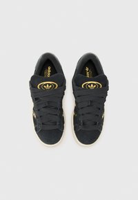 Melni zamšaini sporta apavi ar zelta akcentiem, apaļa logo uz mēlītes, plakani auklas un gaiši brūna gumijas zole. Klasisks sportiskais dizains.