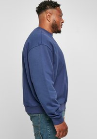 Sötétkék kereknyakú pulóver, laza fit, bordás mandzsettákkal és hempergő anyaggal. Világoskék denim nadrággal viselve.
