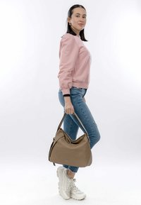 Brown textured handbag with a slouchy design and short straps, held in hand. Worn with light blue jeans and a pink sweatshirt.