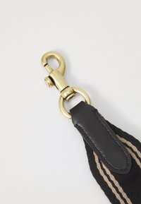 Clip en métal doré attaché à une sangle en tissu noir avec des accents marron, présentant une finition en cuir lisse et une texture tissée.