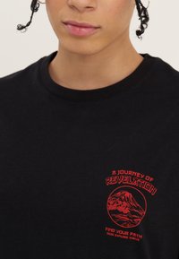 Nahaufnahme einer Person, die ein schwarzes Shirt mit rotem Bergmotiv und dem Text „Eine Reise der Offenbarung, Finde deinen Weg, Suche, erkunde, gedeihe.“ trägt.