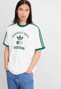White cotton t-shirt with green ribbed collar and sleeves, featuring "ORIGINALS DEPT." logo and Adidas branding, styled with relaxed-fit jeans.