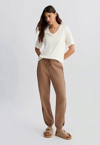 Donna in piedi con le mani dietro la schiena, indossa una maglietta bianca a maniche corte con scollatura a V, pantaloni jogger marroni con cordino e sandali intrecciati beige.