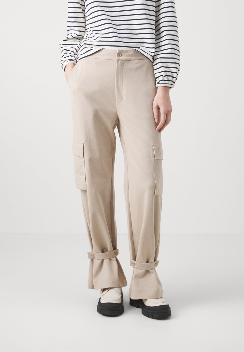 Pantalon cargo beige avec des brides boutonnées au niveau des chevilles, porté avec des baskets blanches et noires à semelles épaisses et un haut blanc à manches longues avec des rayures noires.