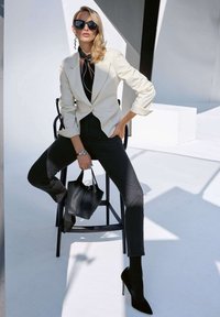 Cremefarbener Blazer mit Dreiviertelärmeln, schwarzes Oberteil, taillierte schwarze Hose, schwarze Stilettos und eine schwarze Handtasche. Sonnenbrille mit großen Rahmen.