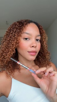 Un crayon cosmétique transparent avec une étiquette colorée, tenu par une personne aux cheveux bouclés. Le crayon présente un design argenté et bleu, ainsi qu'une texture lisse.