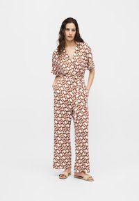 Γυναίκα που φοράει ένα καφέ και λευκό ολόσωμο με γεωμετρικό σχέδιο και κοντά μανίκια, στέκεται με τα χέρια στις τσέπες και μπεζ σανδάλια.