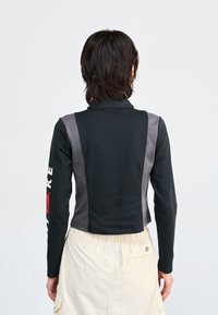 Top negru cu maneci lungi, cu panouri laterale gri și guler înalt. Are text alb pe mâneca stângă, design scurtat și textură netedă.