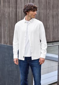 Weißes Hemd mit Knöpfen aus leichtem Stoff, kombiniert mit einem schlichten weißen T-Shirt und dunkelblauen Jeans, steht vor einem Holz-Hintergrund.